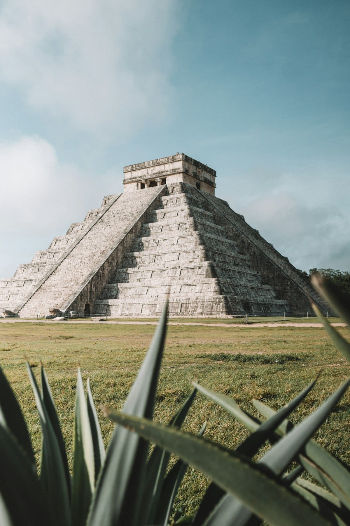 El Castillo, a large step pyramid at Chichen Itza in Mexico, stands under a blue sky with green grass and plants in the foreground.