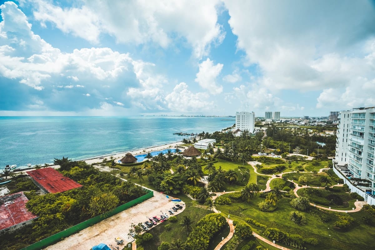 Aerial view of a coastal city with high-rise buildings, a beach, green park areas, and a cloudy sky over the ocean.